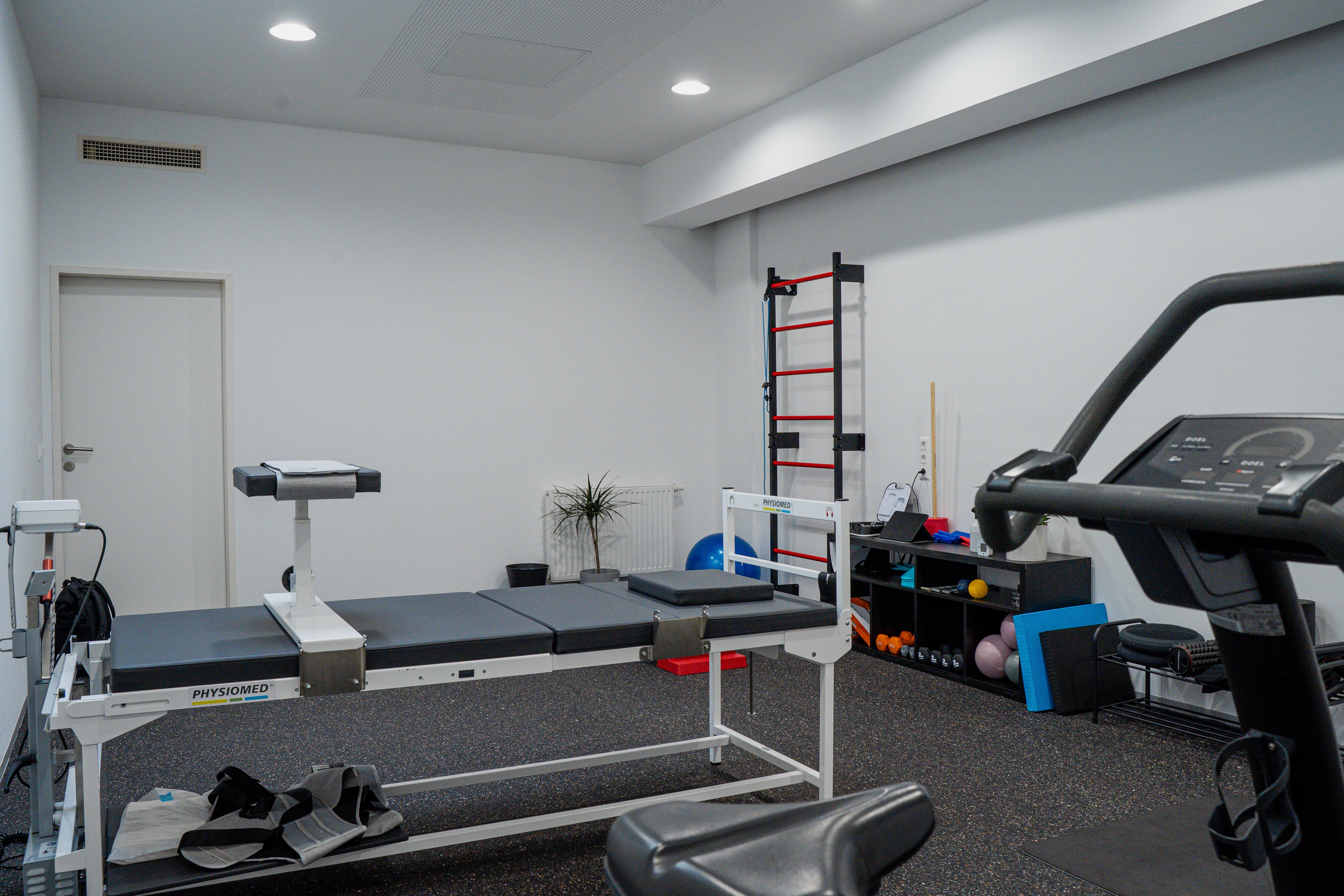 Modern physiotherapy room with a treatment table, treadmill, and wall-mounted exercise equipment.