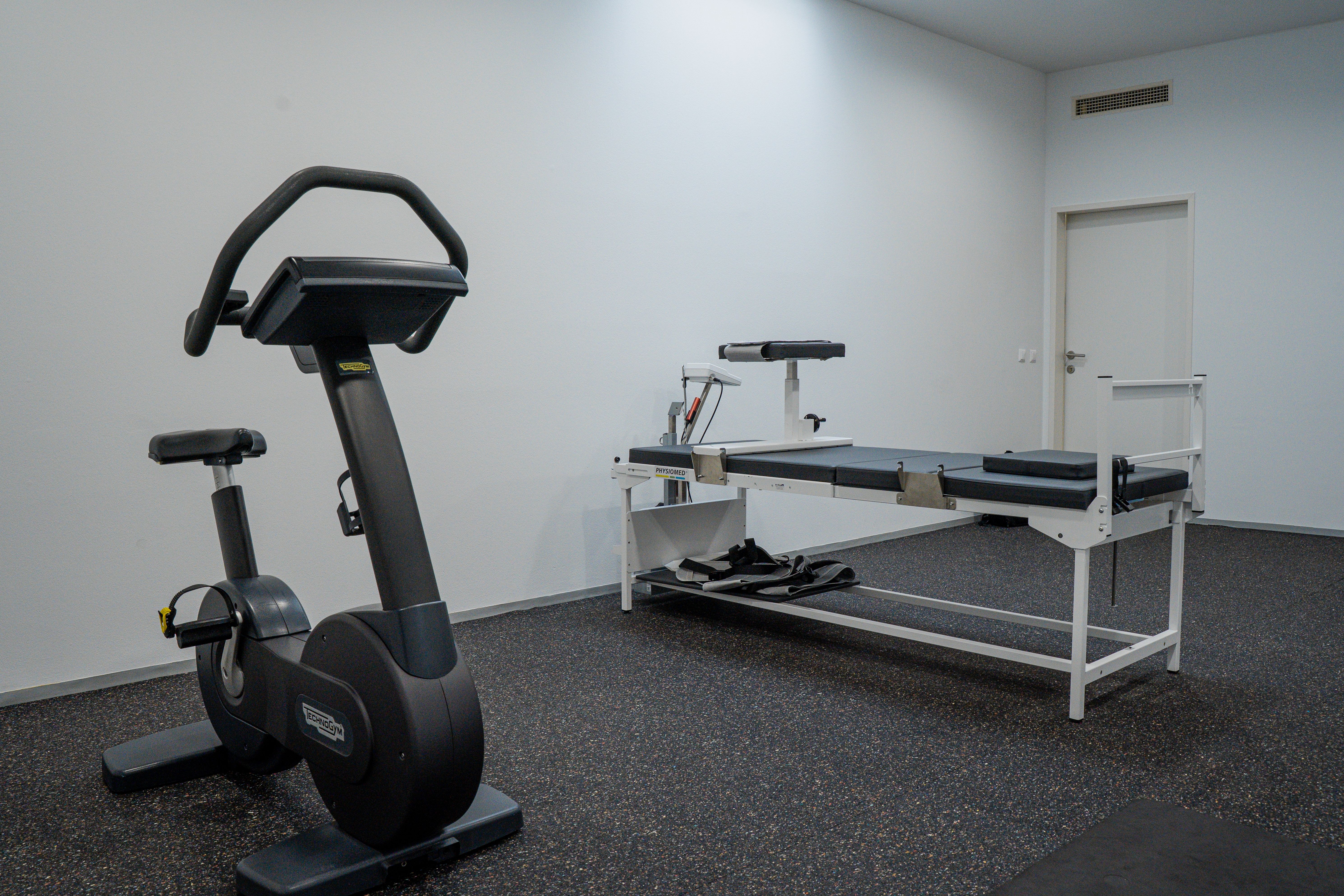 Stationary exercise bike and physical therapy table in a clean, minimalist rehabilitation room.